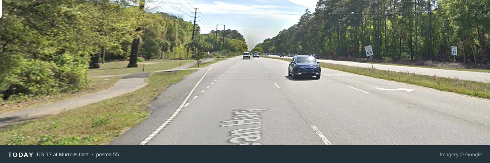 US Highway 17 at Murrells Inlet today, photographed from the roadway. The walking path is a narrow strip on the far left, separated from four lanes of traffic by grass. A speed limit sign reading 55 stands beyond the far side of the road.